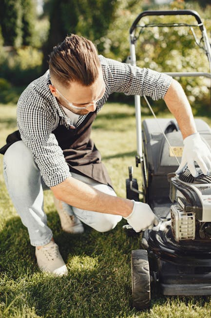 A man wearing safety glasses, a checked shirt, and white gloves is kneeling on a well-maintained lawn, inspecting a black lawnmower. The lawn features neatly trimmed grass, garden beds with a variety
