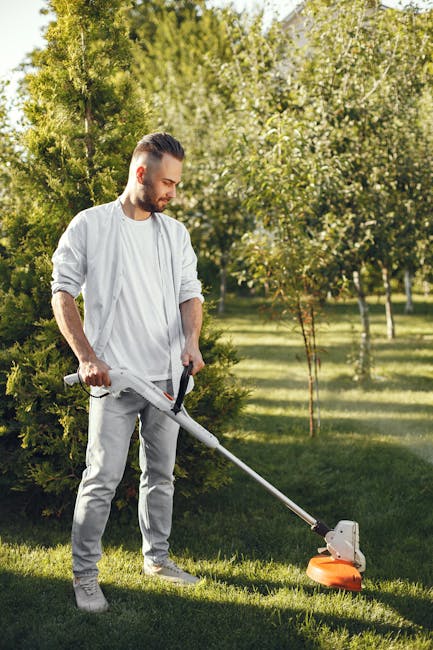 A man wearing safety glasses, a checked shirt, and white gloves is kneeling on a well-maintained lawn, inspecting a black lawnmower. The lawn features neatly trimmed grass, garden beds with a variety